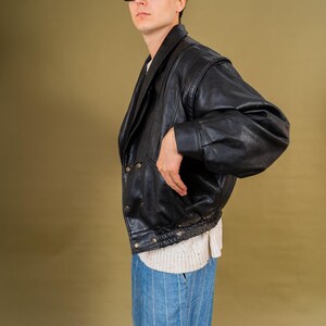 May include: A black leather jacket with a double-breasted design and a ribbed hem. The jacket has a wide collar and is worn over a white sweater and blue striped pants. The person is wearing black sunglasses.