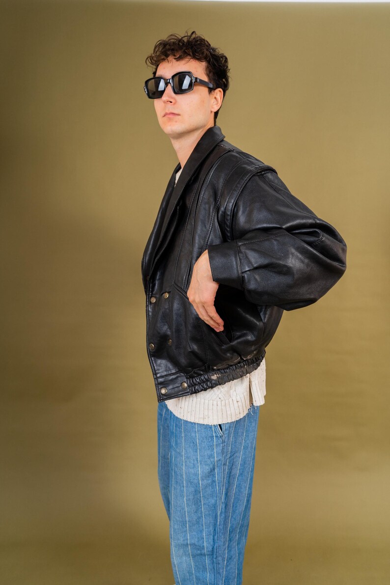 May include: A black leather jacket with a double-breasted design and a ribbed hem. The jacket has a wide collar and is worn over a white sweater and blue striped pants. The person is wearing black sunglasses.