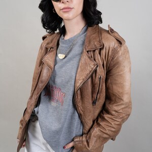 May include: A woman wearing a brown leather jacket, a grey sweatshirt with a red graphic, white shorts, and a brown baseball cap with a white "NY" logo. She is also wearing sunglasses and a gold necklace.