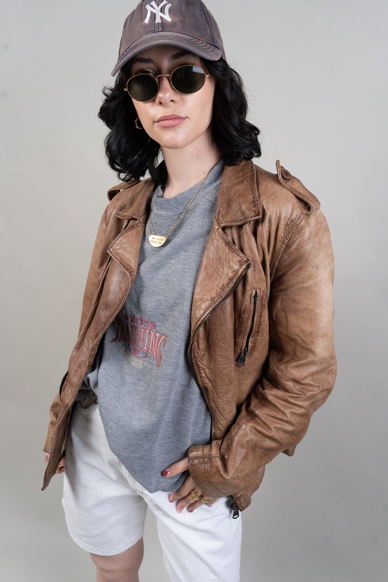May include: A woman wearing a brown leather jacket, a grey sweatshirt with a red graphic, white shorts, and a brown baseball cap with a white "NY" logo. She is also wearing sunglasses and a gold necklace.