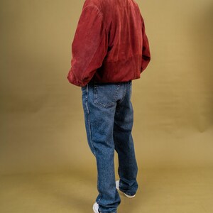 May include: A person wearing a burgundy suede jacket with a brown collar, blue jeans, and white sneakers. The jacket has a relaxed fit. The jeans are a medium wash denim. The background is a solid olive green.