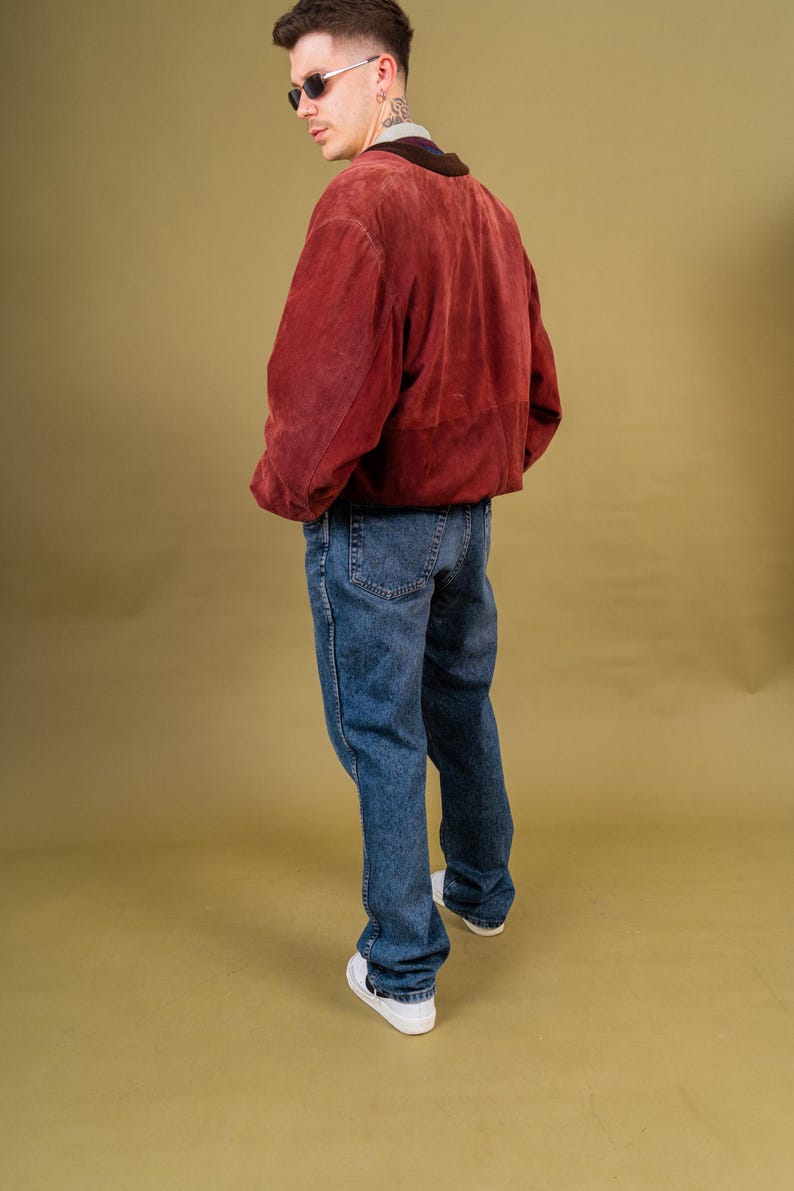 May include: A person wearing a burgundy suede jacket with a brown collar, blue jeans, and white sneakers. The jacket has a relaxed fit. The jeans are a medium wash denim. The background is a solid olive green.