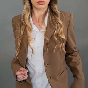 Könnte beinhalten: Eine Frau trägt einen braunen Blazer über einem weißen Hemd mit Knöpfen. Der Blazer hat einen klassischen Stil mit einem Revers und drei Knöpfen.