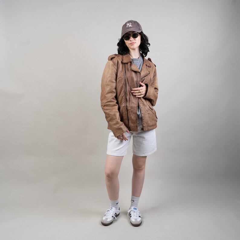 May include: A woman wearing a brown leather jacket, white shorts, and a baseball cap with the New York Yankees logo. She is wearing sunglasses and white trainers.