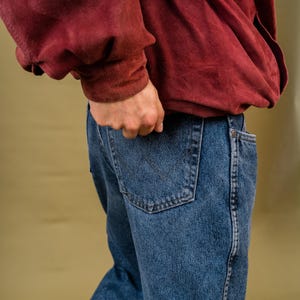 May include: Blue denim jeans with a classic five-pocket design. The jeans have a straight leg cut and a dark wash. A burgundy-colored sweatshirt is partially visible. The person's hand is in the back pocket.