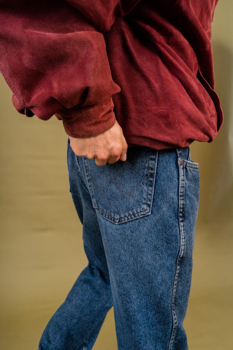 May include: Blue denim jeans with a classic five-pocket design. The jeans have a straight leg cut and a dark wash. A burgundy-colored sweatshirt is partially visible. The person's hand is in the back pocket.