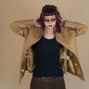 Pode incluir: Uma jaqueta de couro marrom com fecho de z&iacute;per. A jaqueta est&aacute; aberta e o forro &eacute; dourado. A pessoa que est&aacute; usando a jaqueta tem cabelo roxo e est&aacute; usando um top preto sem mangas e cal&ccedil;as de veludo cotel&ecirc; marrons.