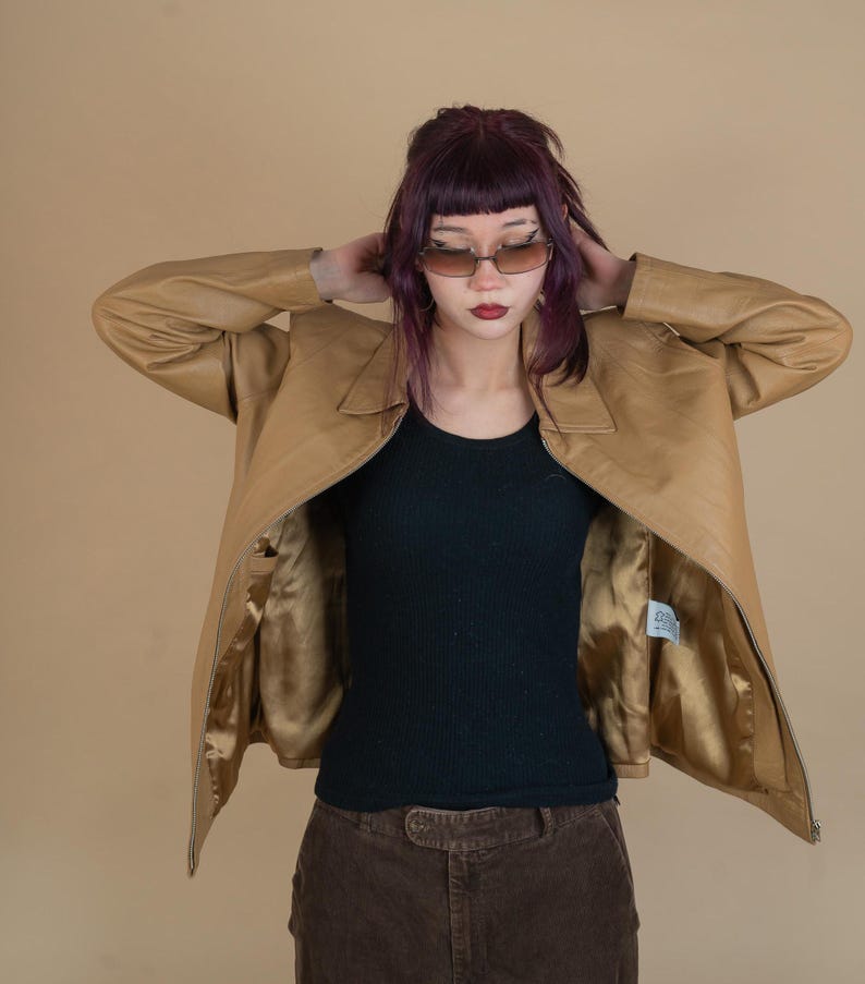 Pode incluir: Uma jaqueta de couro marrom com fecho de z&iacute;per. A jaqueta est&aacute; aberta e o forro &eacute; dourado. A pessoa que est&aacute; usando a jaqueta tem cabelo roxo e est&aacute; usando um top preto sem mangas e cal&ccedil;as de veludo cotel&ecirc; marrons.