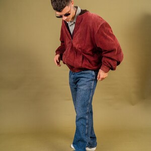 May include: A person wearing a burgundy suede bomber jacket, a gray collared shirt, and blue jeans. The person is also wearing sunglasses and white sneakers with black accents.