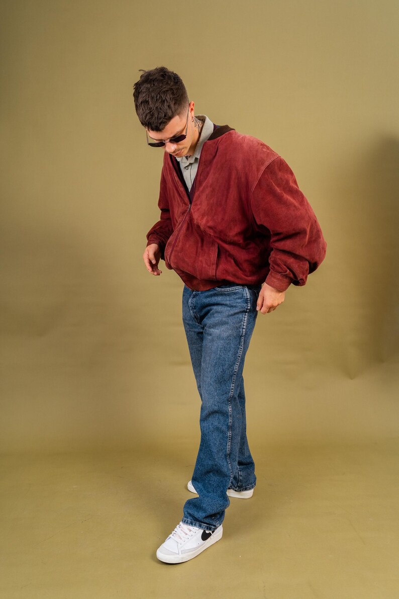 May include: A person wearing a burgundy suede bomber jacket, a gray collared shirt, and blue jeans. The person is also wearing sunglasses and white sneakers with black accents.