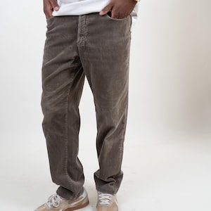 Pode incluir: Um par de calças de veludo cotelê castanhas com um design clássico de cinco bolsos, fecho de botões e corte de perna reta, feito de tecido canelado. As calças são usadas com uma t-shirt branca e sapatilhas bege com solas de borracha.