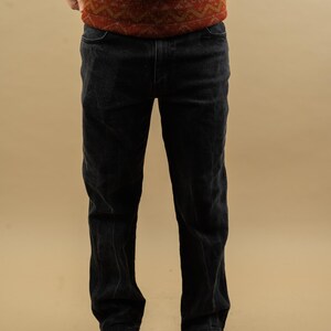 May include: A pair of black denim jeans with a straight leg fit. The jeans are worn by a person wearing a red and brown patterned sweater.