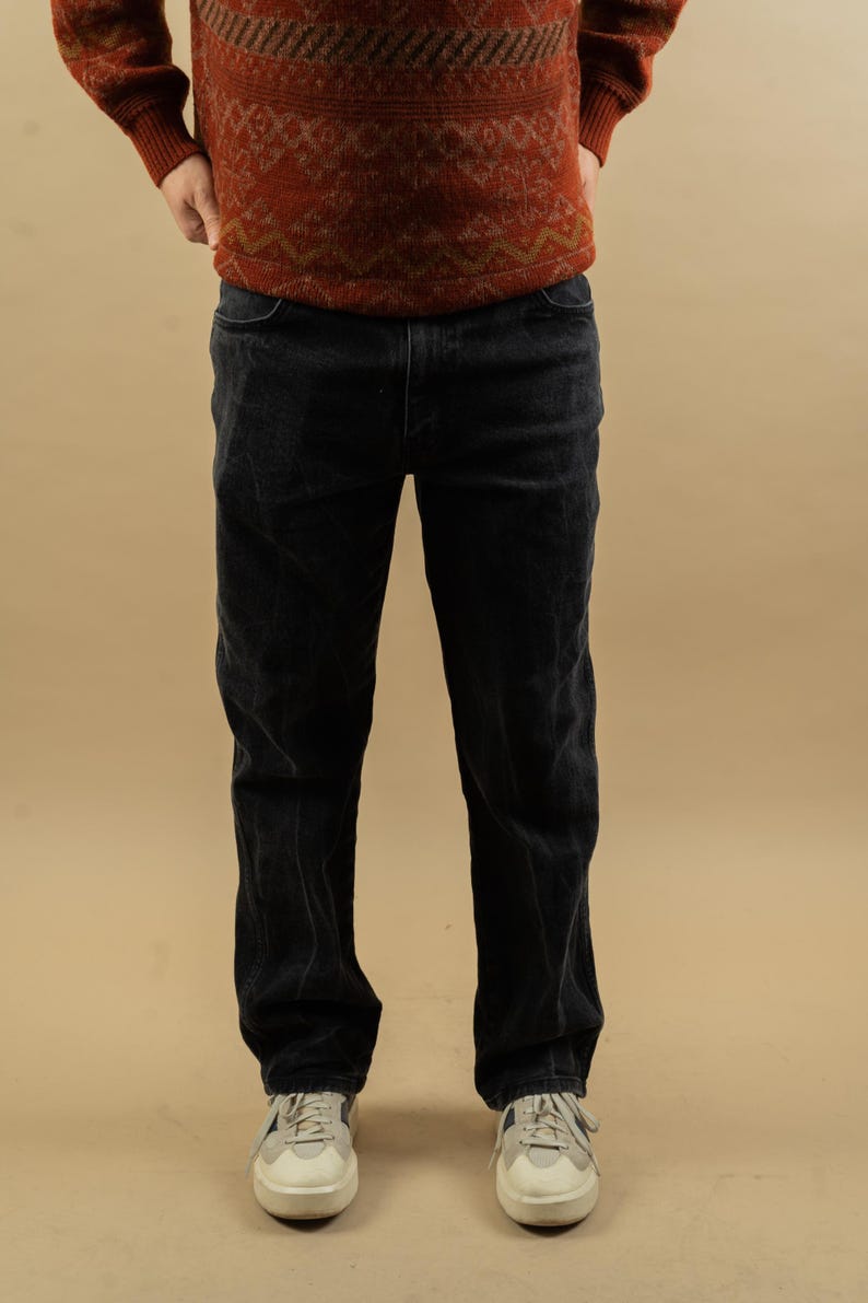 May include: A pair of black denim jeans with a straight leg fit. The jeans are worn by a person wearing a red and brown patterned sweater.