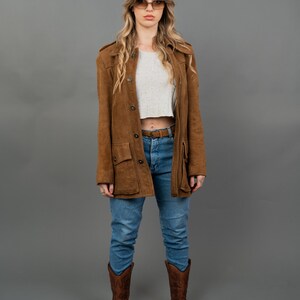 Peut inclure: Veste en daim marron &agrave; boutons, port&eacute;e sur un crop top blanc et un jean bleu. La tenue est compl&eacute;t&eacute;e par des bottes de cowboy marron. La veste a des poches et un col.