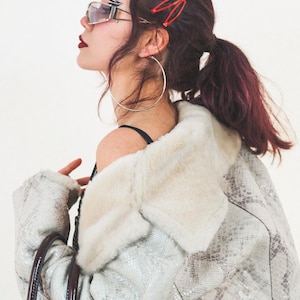 Puede incluir: Una persona con gafas de sol plateadas, pendientes de aro plateados grandes y una pinza para el pelo roja. Lleva una chaqueta forrada de piel sintética con estampado de piel de serpiente y un bolso de mano de cuero marrón. La persona tiene el pelo oscuro y lleva pintalabios oscuro.