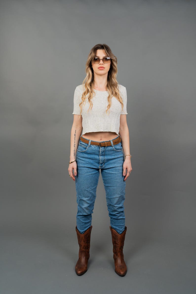 Peut inclure: Une femme porte un haut court blanc &agrave; manches courtes, un jean bleu, une ceinture marron et des bottes de cowboy marron. Elle porte &eacute;galement des lunettes de soleil. Le fond est gris uni.