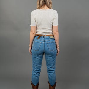 Peut inclure: Une personne porte un haut blanc mouchet&eacute; &agrave; manches courtes, un jean bleu, une ceinture marron et des bottes de cowboy marron. La tenue est pos&eacute;e sur un fond gris.