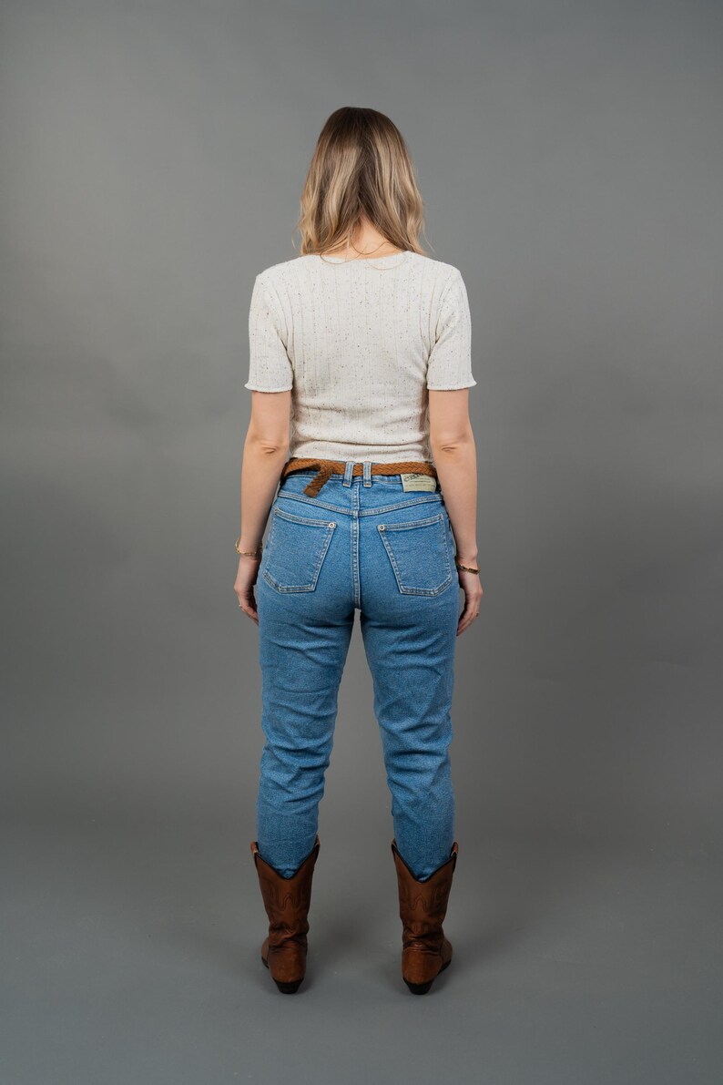 Peut inclure: Une personne porte un haut blanc mouchet&eacute; &agrave; manches courtes, un jean bleu, une ceinture marron et des bottes de cowboy marron. La tenue est pos&eacute;e sur un fond gris.