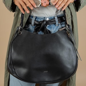 May include: A black leather handbag with a silver buckle and the word "JOOP!" printed on the front. The bag is worn by a person wearing a green jacket and blue jeans.