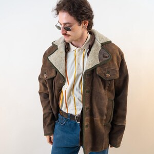 Op de afbeelding: Een bruine su&egrave;de jas met een cr&egrave;me kleurige schapenvacht kraag en afwerking. De jas heeft twee voorzakken en een knoopsluiting. De persoon draagt een gestreepte poloshirt en een blauwe jeans. De jas is een vintage stijl.