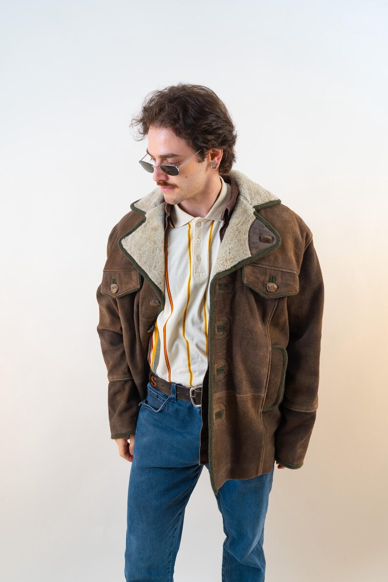 Op de afbeelding: Een bruine su&egrave;de jas met een cr&egrave;me kleurige schapenvacht kraag en afwerking. De jas heeft twee voorzakken en een knoopsluiting. De persoon draagt een gestreepte poloshirt en een blauwe jeans. De jas is een vintage stijl.