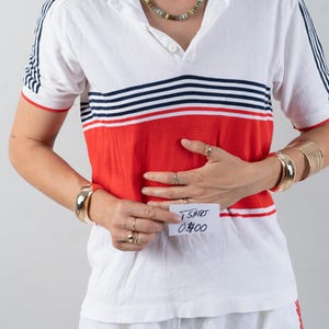 May include: A white and red striped short-sleeved shirt with a white collar and a button-up front. The shirt has a red band across the chest with blue and white stripes above it. The person is wearing a gold bracelet and a gold necklace. A white sticky note with the text "T-SHIRT 0400" is attached to the shirt.
