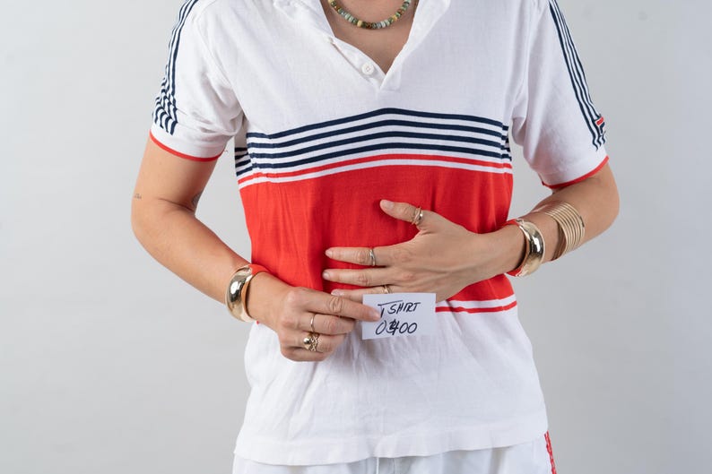 May include: A white and red striped short-sleeved shirt with a white collar and a button-up front. The shirt has a red band across the chest with blue and white stripes above it. The person is wearing a gold bracelet and a gold necklace. A white sticky note with the text "T-SHIRT 0400" is attached to the shirt.