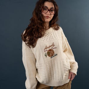 Puede incluir: Un suéter de punto de cable blanco con un parche bordado de colores que dice "Aropole Flew of Ducks in a line".