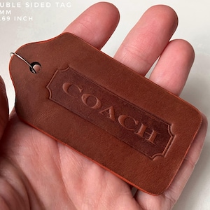 Op de afbeelding: Een bruin lederen label met het woord "COACH" in reliëf in een decoratief kader. Het label is 87 mm bij 43 mm en heeft een metalen lus.