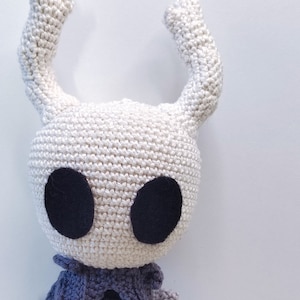 May include: A handmade crochet doll featuring a character with a white head, large black eyes, and two curved horns. The doll has a dark blue cape and a black body, showcasing intricate stitching and a whimsical design. The doll is approximately 6 inches tall.