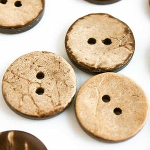 May include: Close-up of several round, natural coconut buttons with two or three holes each. The buttons have a light tan center and a dark brown edge. The buttons are scattered on a white surface.