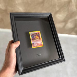 Könnte beinhalten: Eine gerahmte Charizard Pokémon-Sammelkarte. Die Karte ist auf einem schwarzen Hintergrund in einem schwarzen Rahmen zentriert. Die Karte hat einen gelben Rand mit einer Charizard-Illustration und Text. Der Rahmen hat unten den Text "1/100".