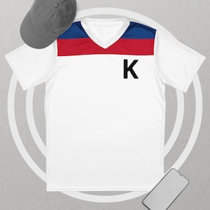 Könnte beinhalten: Weißes V-Ausschnitt-Sportshirt mit blau-rotem Querstreifen auf der Brust. Auf der Vorderseite ist ein großer schwarzer Buchstabe "K" aufgedruckt. Eine graue Baseballkappe ist oben links zu sehen. Ein Smartphone befindet sich unten rechts.