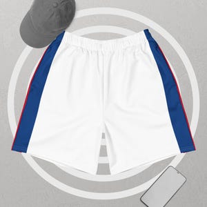 Op de afbeelding: Witte sportbroek met blauwe en rode zijstrepen. De shorts hebben een elastische tailleband en een relaxte pasvorm. Een grijze baseballcap is linksboven zichtbaar. Een smartphone staat rechtsonder.