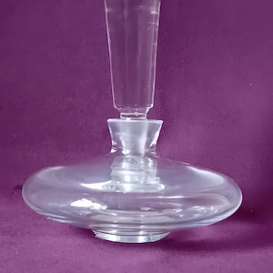 May include: A clear glass decanter featuring a wide, flattened circular base and a tall, slender, cylindrical stopper that fits securely into the narrow neck. The vessel is constructed from transparent glass with a smooth, reflective surface.