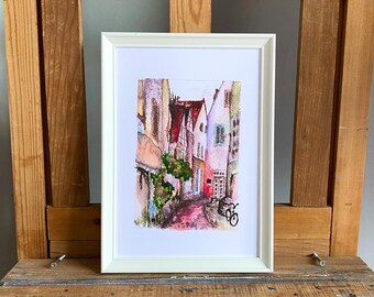 May include: A framed watercolor painting of a European street scene. The artwork features colorful buildings with red-tiled roofs, a cobblestone street, and a bicycle. The painting is set in a white frame and rests on a wooden surface.