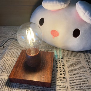 May include: A wooden lamp base with a light bulb and a power button. The lamp is on a patterned surface with a white plush toy in the background.