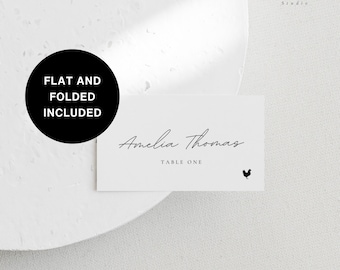 May include: White place card with black text that reads "Amelia Thomas" and "Table One". A small black rooster icon is in the bottom right corner. The place card is on a white surface.