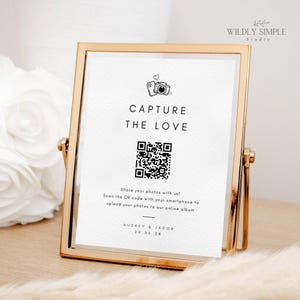 Könnte beinhalten: Ein weißes Schild in einem goldenen Rahmen mit einem schwarz-weißen QR-Code. Das Schild lautet "Capture the Love" und enthält Anweisungen, den QR-Code mit einem Smartphone zu scannen, um Fotos in ein Online-Album hochzuladen. Das Schild enthält auch die Namen "Audrey & Jacob" und das Datum "20.05.28".