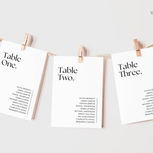May include: Three white cards with black text are clipped to a string. Each card reads "Table One," "Table Two," and "Table Three." Below each table number are names. The cards are for a wedding or event.