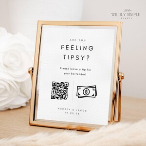 May include: A white sign with black text and graphics in a gold frame. The sign reads "Are you feeling tipsy? Please leave a tip for your bartender!" with a QR code and a graphic of cash. The sign also includes the text "Audrey & Jacob 20.05.28".