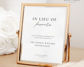May include: A gold-framed sign with the text "IN LIEU OF favours" and a message stating that a donation has been made to The Animal Welfare Foundation in lieu of wedding favors. The sign also includes the names Cecilia & Thomas and the date 17.05.2025.