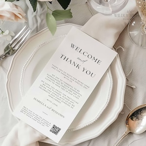 May include: A white wedding menu card with the text "WELCOME and THANK YOU" in black script, placed on a white plate with a silver rim. The card includes a message and a QR code. A silver fork and spoon are visible.