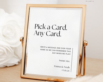 May include: A gold-framed sign with the text "Pick a Card. Any Card." and additional text inviting guests to write a message and sign their name. The sign includes the names "Emma & Noah" and the date "17.05.25".