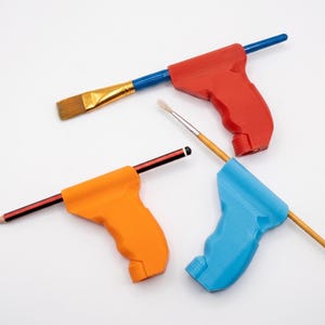 May include: Three ergonomic pistol-grip style holders in red, orange, and blue, each fitted with a different writing or painting tool. The red holder contains a flat-tipped paintbrush, the orange holds a pencil, and the blue holds a fine-tipped brush.