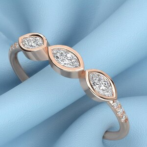 Op de afbeelding: Ros&eacute;gouden ring met drie marquise-geslepen diamanten en kleine accentstenen. De ring heeft een delicate band en een gepolijste afwerking, tegen een lichtblauwe achtergrond. Een sieraad voor dagelijks gebruik of speciale gelegenheden.