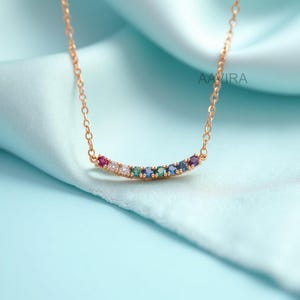 May include: A gold-colored necklace with a curved bar pendant. The pendant is adorned with a row of colorful gemstones, including red, white, green, blue, and purple. The necklace is displayed on a light blue fabric.