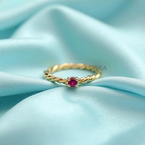 May include: A gold-colored ring with a twisted band design, featuring a small, round, red gemstone at the center. The ring is set against a light blue fabric background. The text "AAVIRA" is visible on the ring.