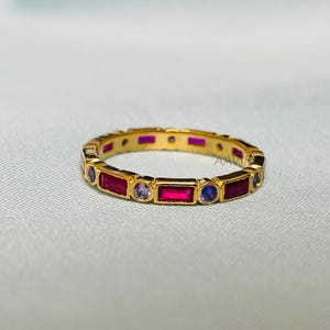 May include: A gold band ring featuring a row of alternating red and blue rectangular gemstones. The ring is displayed on a light blue fabric background.