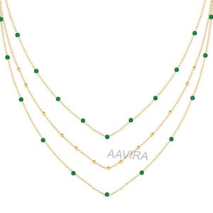 May include: A gold chain necklace with three strands of delicate chain and small green gemstones. The necklace is layered and features a subtle, elegant design.